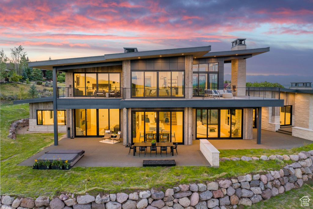 Back house at dusk featuring a balcony, a yard, and a patio