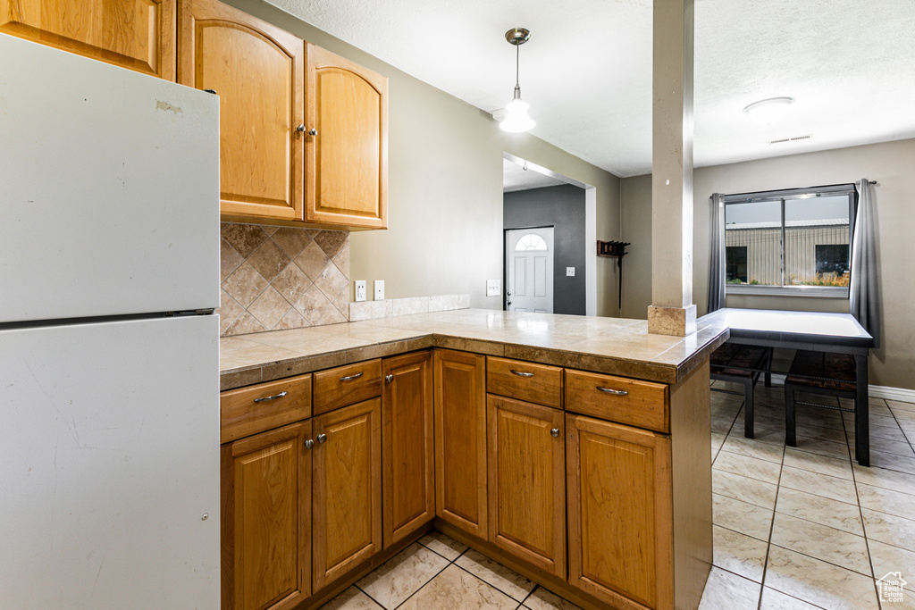 Kitchen with light tile patterned floors, white refrigerator, tasteful backsplash, hanging light fixtures, and kitchen peninsula