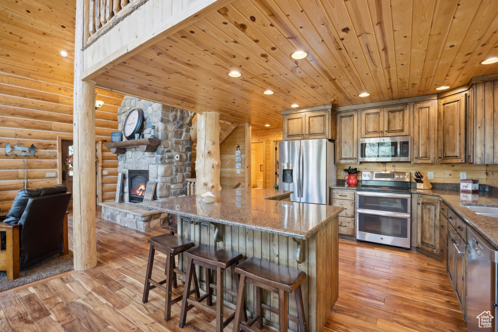 Kitchen featuring light wood-type flooring, rustic walls, stainless steel appliances, wooden ceiling, and a stone fireplace