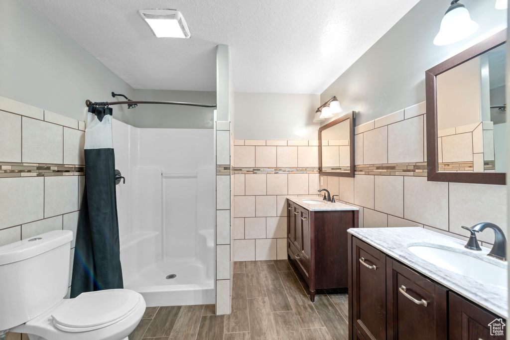 Bathroom with dual vanity, tile walls, a textured ceiling, toilet, and a shower