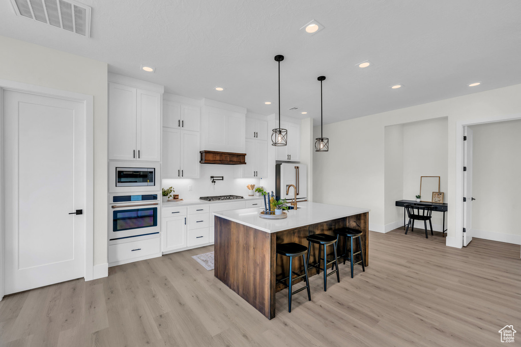 Kitchen featuring white cabinetry, light wood-type flooring, an island with sink, appliances with stainless steel finishes, and pendant lighting