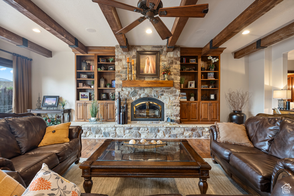 Living room featuring beam ceiling, built in shelves, ceiling fan, a stone fireplace, and wood-type flooring