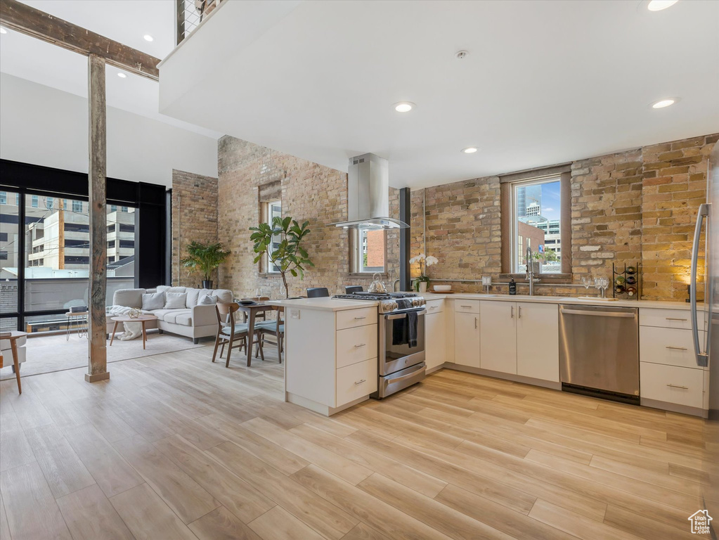 Kitchen featuring appliances with stainless steel finishes, island range hood, kitchen peninsula, light hardwood / wood-style floors, and brick wall