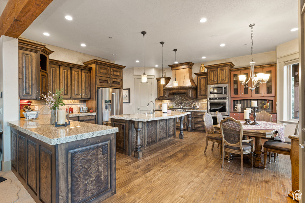 Kitchen with appliances with stainless steel finishes, tasteful backsplash, light stone counters, custom exhaust hood, and a large island with sink