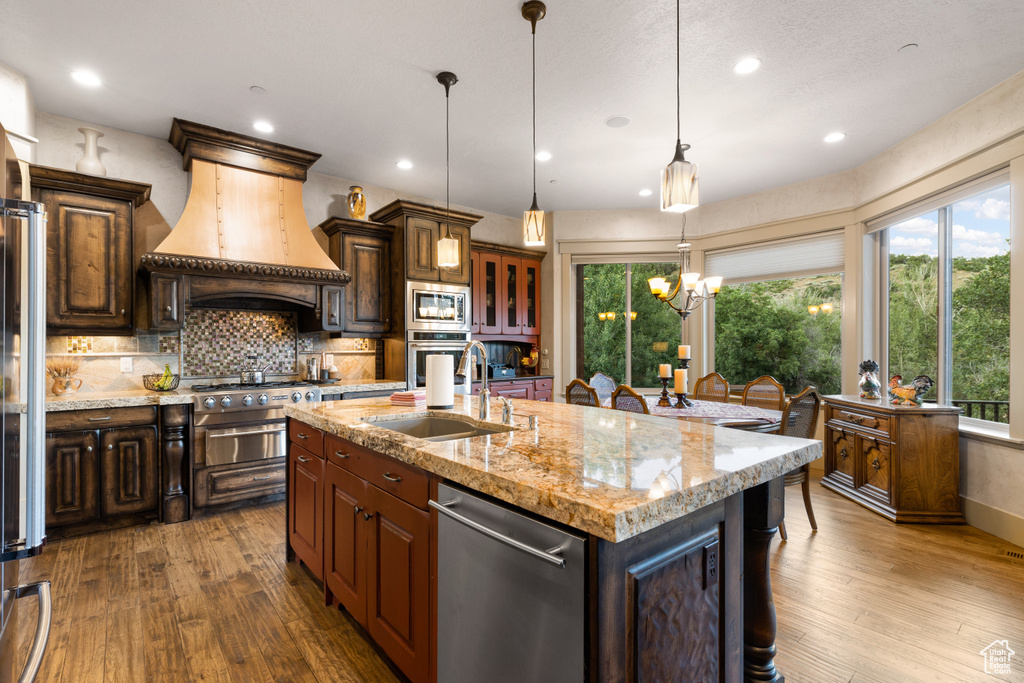 Kitchen with custom exhaust hood, hardwood / wood-style flooring, stainless steel appliances, a center island with sink, and backsplash