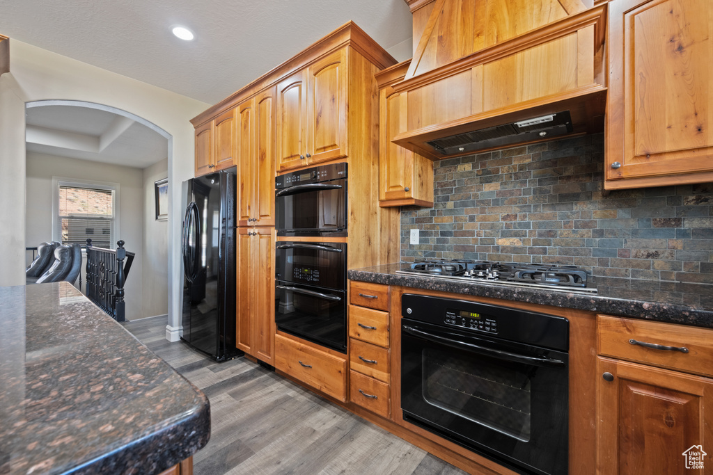 Kitchen featuring custom range hood, dark stone counters, light hardwood / wood-style floors, black appliances, and decorative backsplash