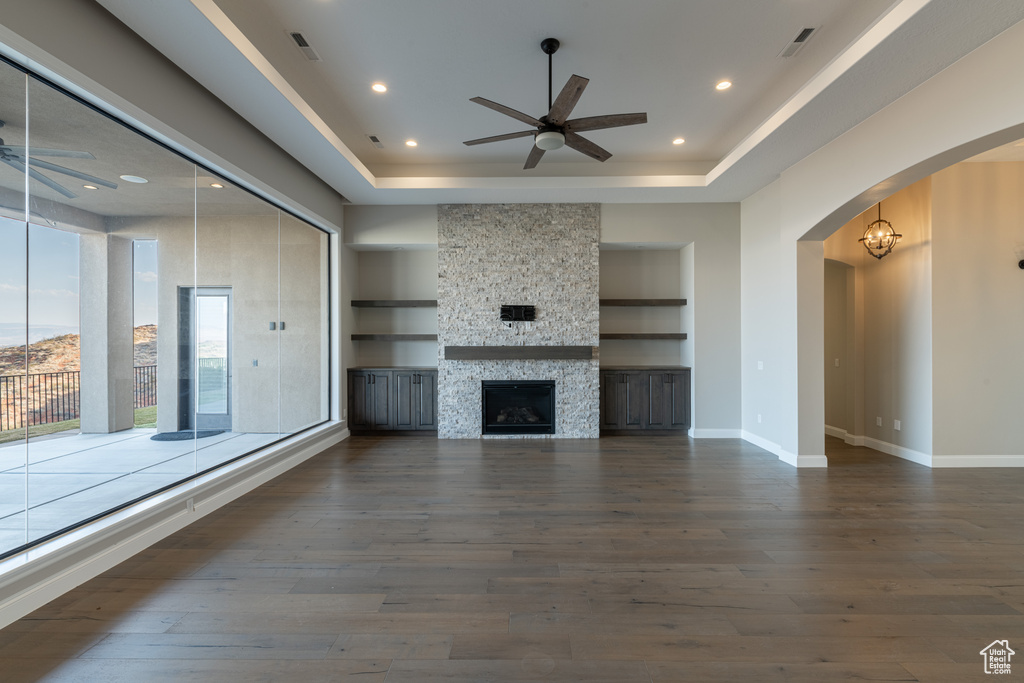 Unfurnished living room featuring a stone fireplace, a raised ceiling, built in features, and ceiling fan