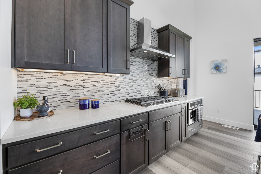 Kitchen featuring dark brown cabinets, stainless steel appliances, light hardwood / wood-style floors, wall chimney exhaust hood, and decorative backsplash