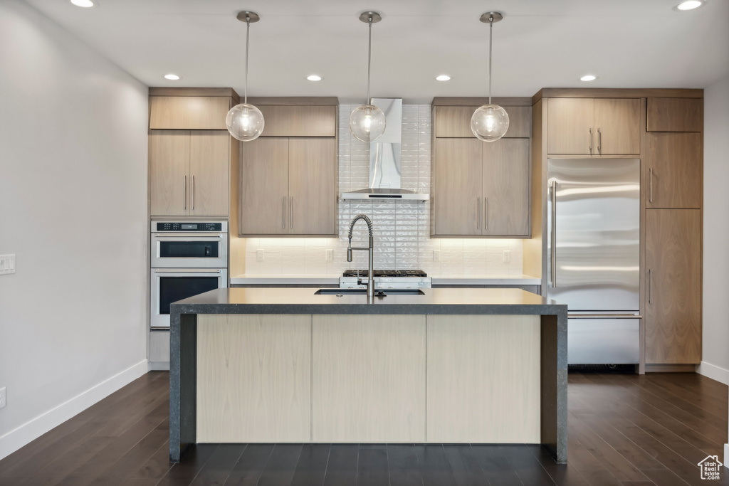 Kitchen with dark hardwood / wood-style flooring, decorative backsplash, wall chimney exhaust hood, appliances with stainless steel finishes, and a kitchen island with sink