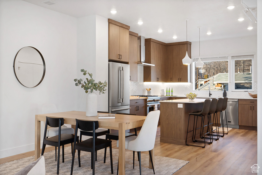 Kitchen with wall chimney range hood, stainless steel appliances, hanging light fixtures, light wood-type flooring, and a center island