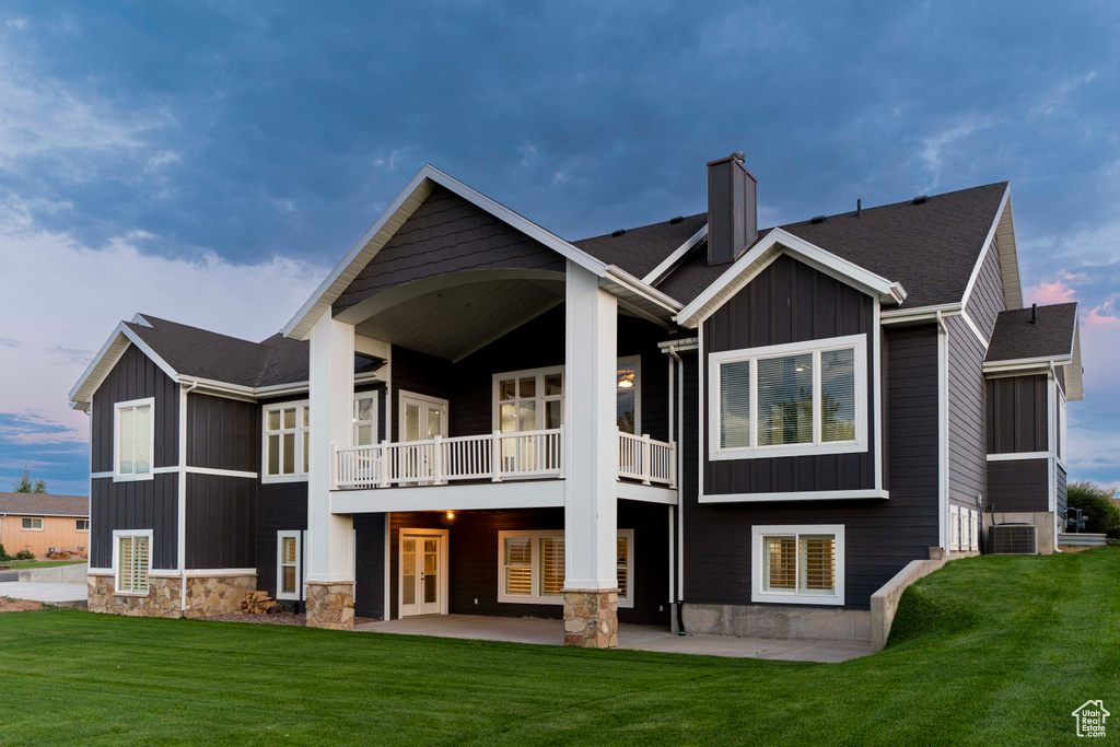 Back house at dusk featuring central air condition unit, a balcony, a patio area, and a lawn