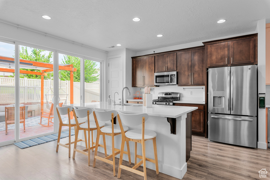 Kitchen featuring light hardwood / wood-style flooring, a kitchen island with sink, dark brown cabinetry, stainless steel appliances, and a breakfast bar area