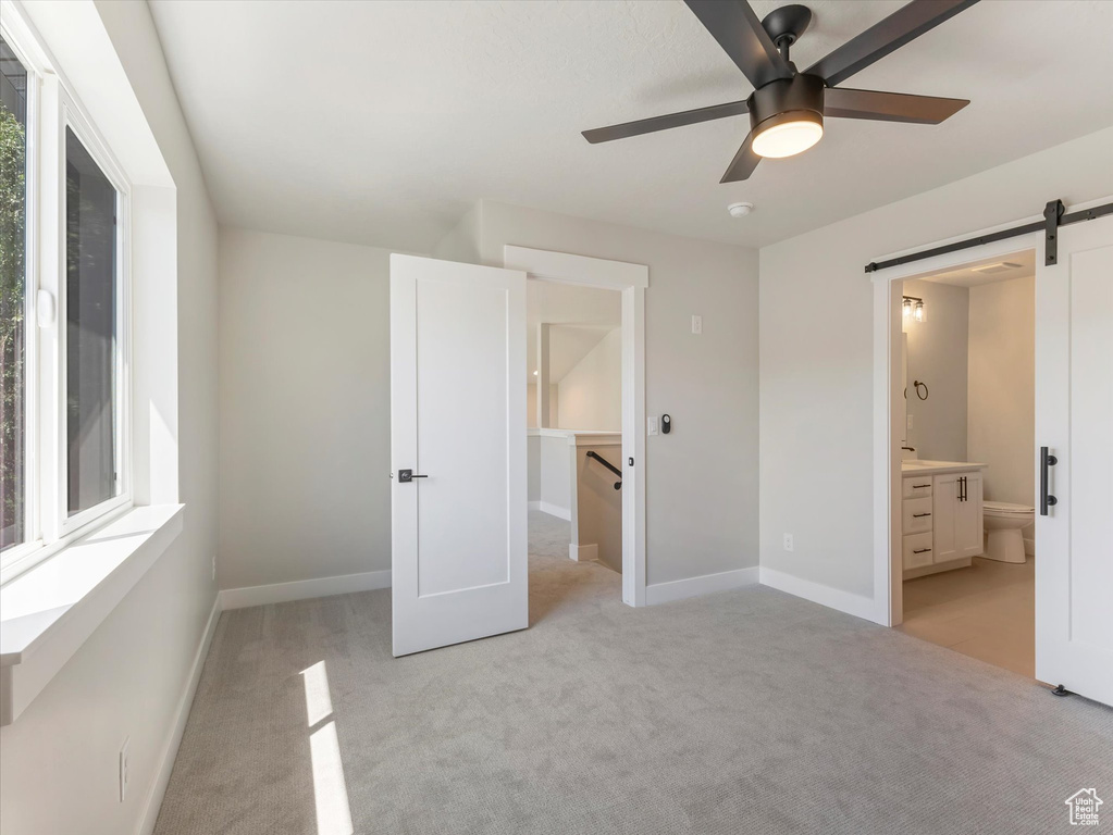 Unfurnished bedroom featuring a barn door, light carpet, ceiling fan, a spacious closet, and ensuite bathroom