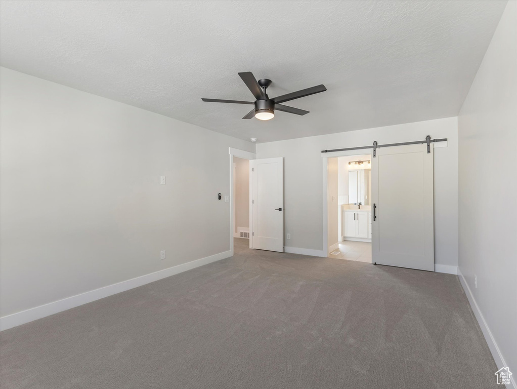 Unfurnished bedroom with ceiling fan, a barn door, light carpet, and connected bathroom