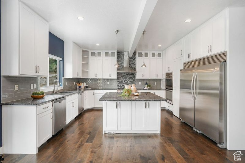 Kitchen with appliances with stainless steel finishes, white cabinets, tasteful backsplash, a center island, and wall chimney range hood