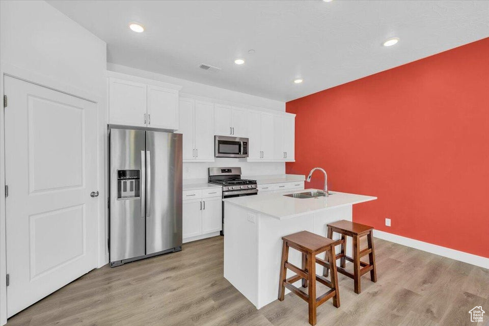 Kitchen with appliances with stainless steel finishes, a kitchen island with sink, sink, light hardwood / wood-style flooring, and white cabinets