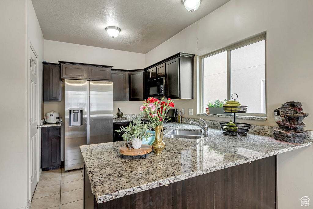 Kitchen featuring high end fridge, light tile patterned flooring, kitchen peninsula, dark brown cabinetry, and light stone counters