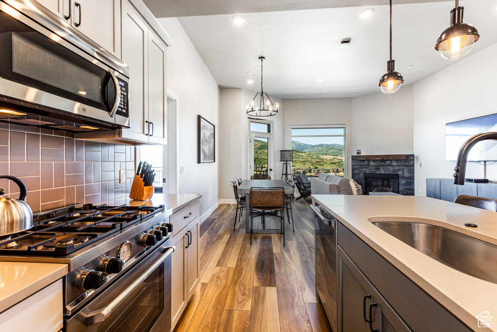 Kitchen with decorative backsplash, appliances with stainless steel finishes, a stone fireplace, pendant lighting, and light hardwood / wood-style floors