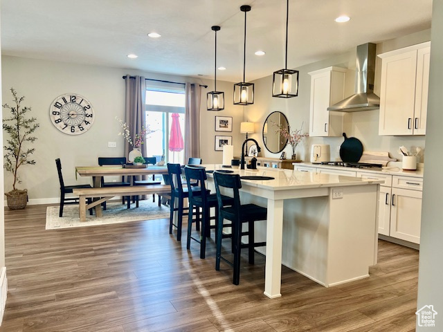 Kitchen with a kitchen island with sink, wall chimney range hood, white cabinets, hardwood / wood-style flooring, and decorative light fixtures