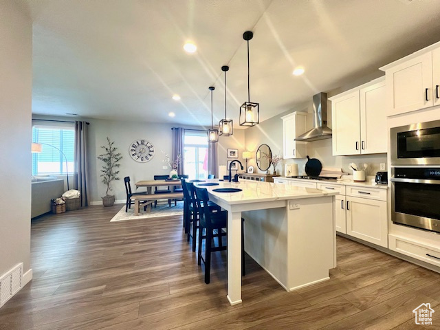 Kitchen featuring wall chimney range hood, an island with sink, dark hardwood / wood-style flooring, and a wealth of natural light