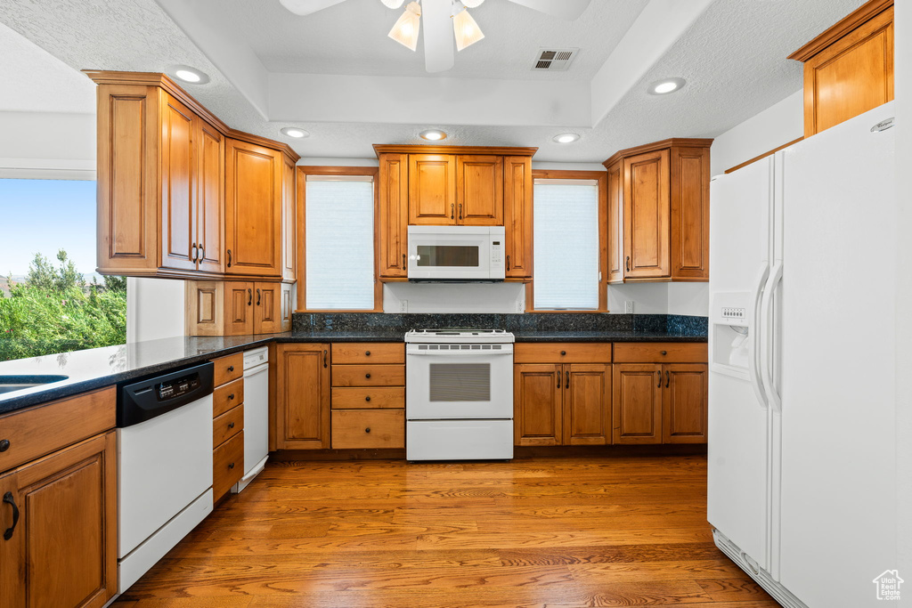 Kitchen featuring white appliances, light wood-type flooring, a tray ceiling, dark stone counters, and ceiling fan