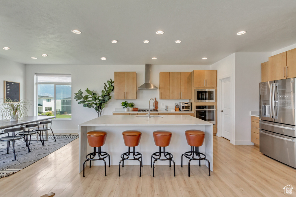 Kitchen with light wood-type flooring, stainless steel appliances, a kitchen island with sink, and wall chimney range hood