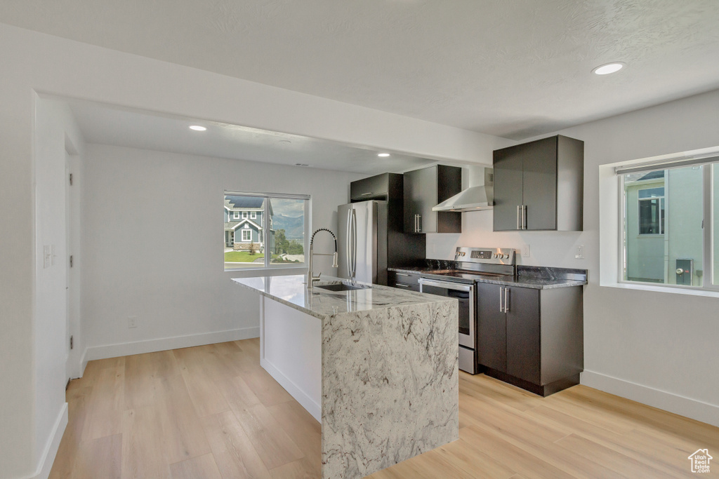 Kitchen featuring wall chimney exhaust hood, light stone countertops, stainless steel appliances, and light hardwood / wood-style floors