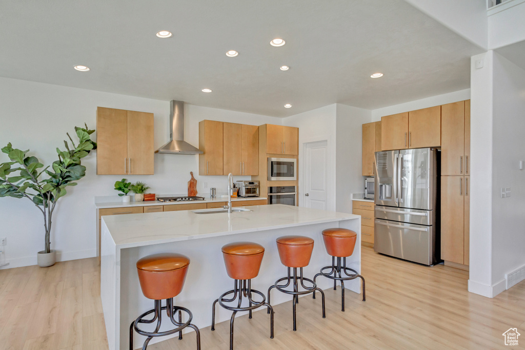 Kitchen featuring stainless steel appliances, light hardwood / wood-style flooring, wall chimney exhaust hood, and a kitchen island with sink