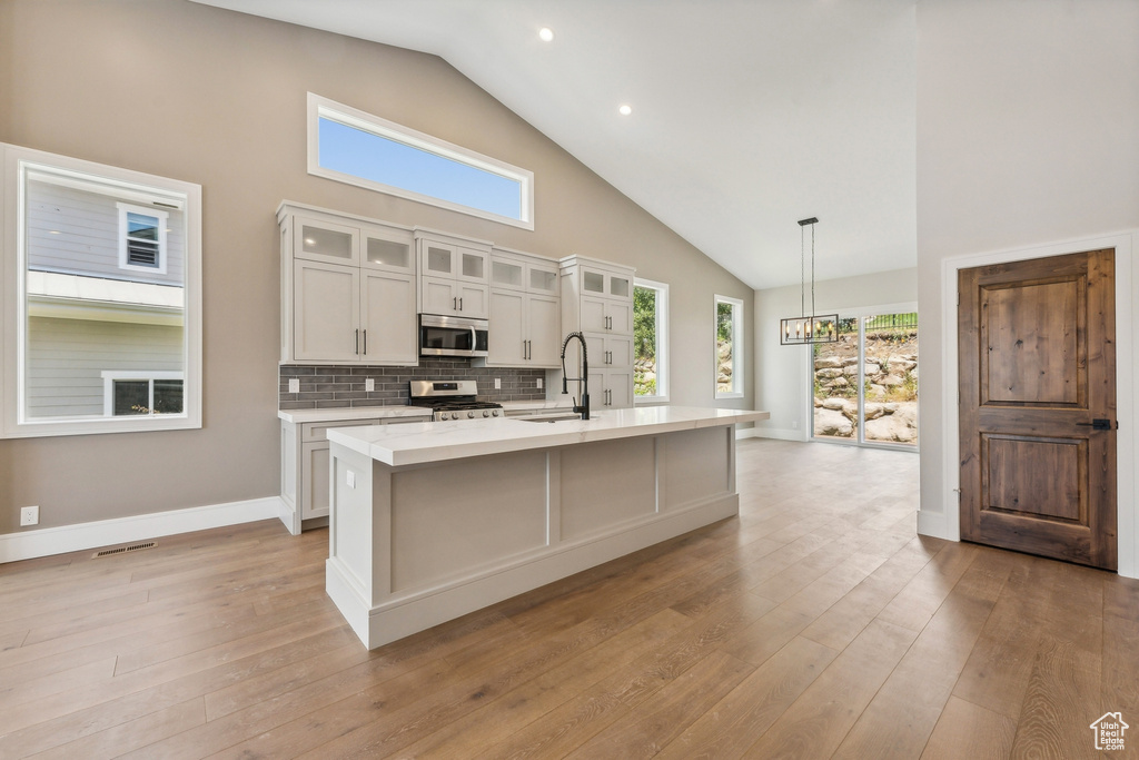 Kitchen featuring appliances with stainless steel finishes, high vaulted ceiling, a center island with sink, tasteful backsplash, and light hardwood / wood-style flooring