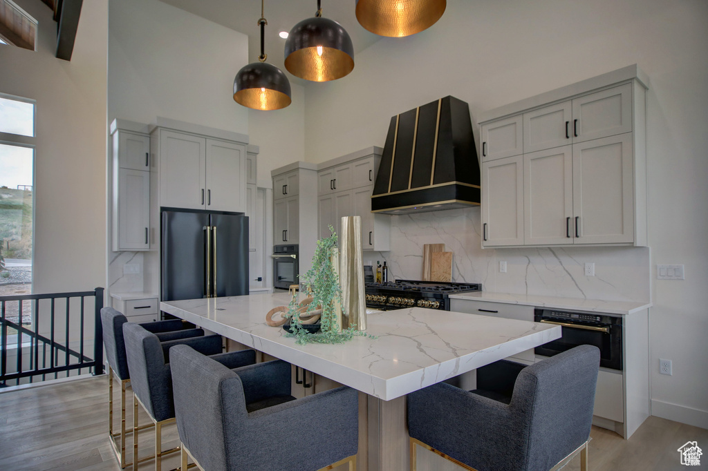 Kitchen featuring a kitchen island with sink, light wood-type flooring, backsplash, black oven, and wall chimney exhaust hood