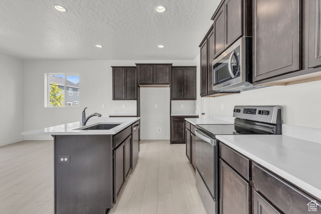 Kitchen with a textured ceiling, an island with sink, stainless steel appliances, sink, and light wood-type flooring