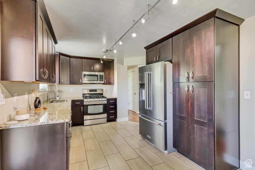 Kitchen with dark brown cabinetry, track lighting, appliances with stainless steel finishes, light stone countertops, and sink