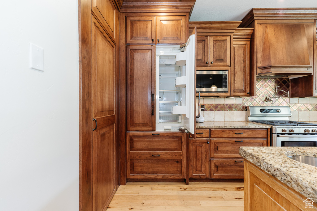 Kitchen with appliances with stainless steel finishes, custom range hood, light hardwood / wood-style floors, decorative backsplash, and light stone countertops