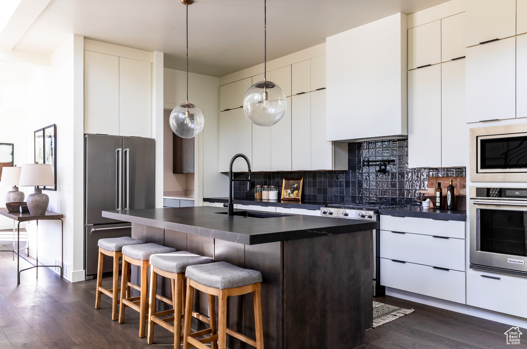 Kitchen featuring stainless steel appliances, a kitchen island with sink, sink, decorative backsplash, and dark wood-type flooring