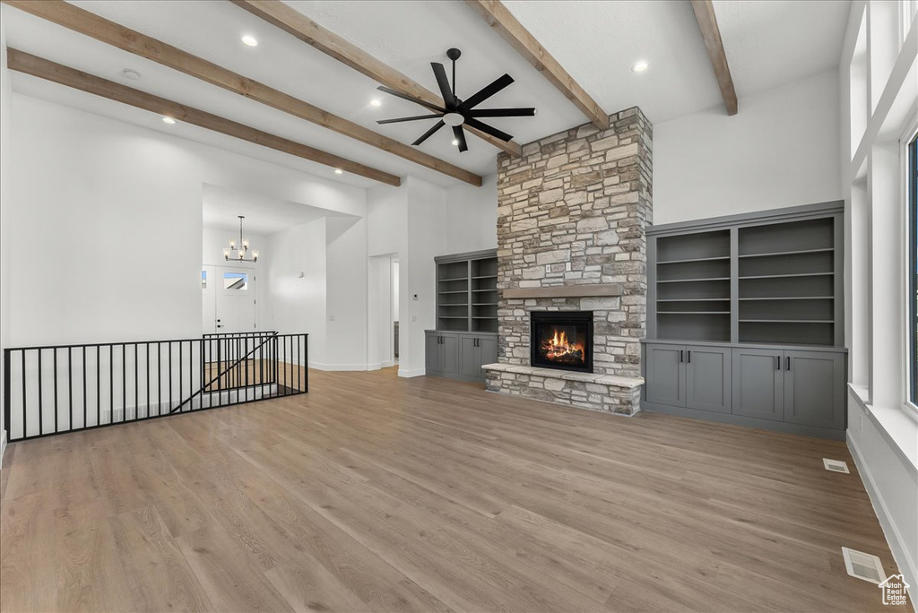 Unfurnished living room with built in shelves, light hardwood / wood-style flooring, a stone fireplace, ceiling fan with notable chandelier, and beam ceiling