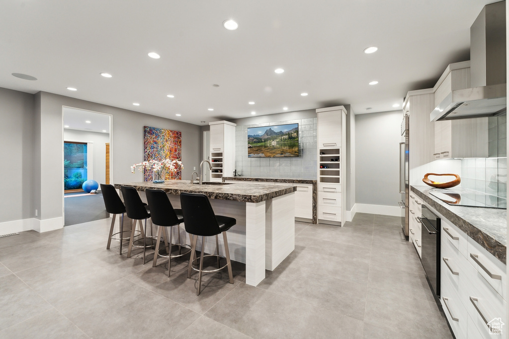 Kitchen featuring backsplash, a large island with sink, wall chimney range hood, and white cabinets