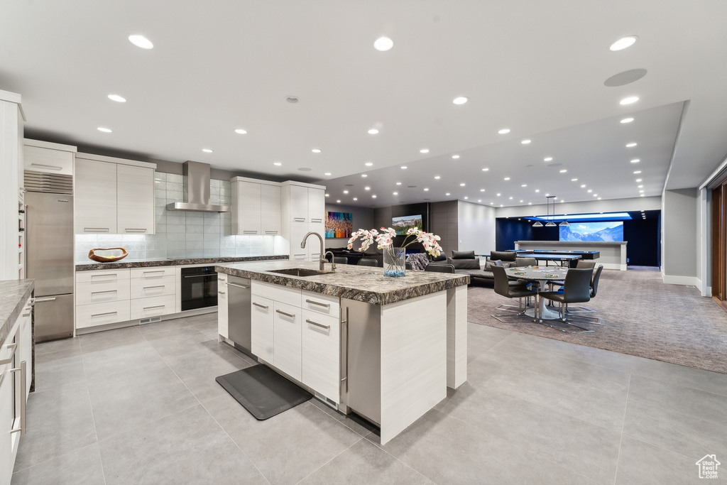 Kitchen with decorative backsplash, white cabinetry, stainless steel appliances, a center island with sink, and wall chimney range hood