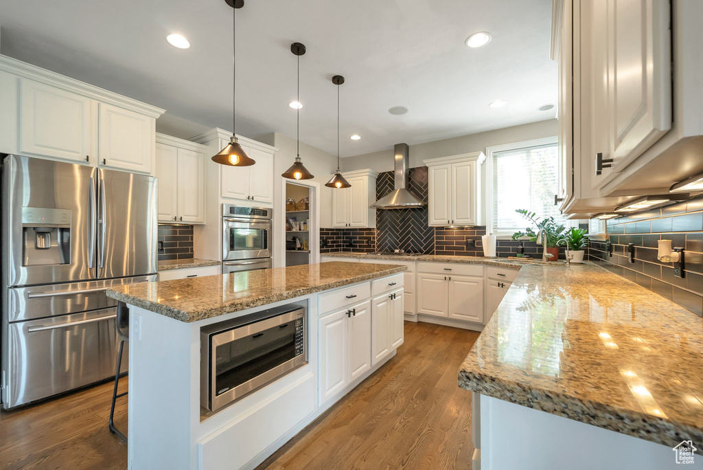 Kitchen featuring appliances with stainless steel finishes, backsplash, white cabinetry, wall chimney range hood, and hardwood / wood-style flooring