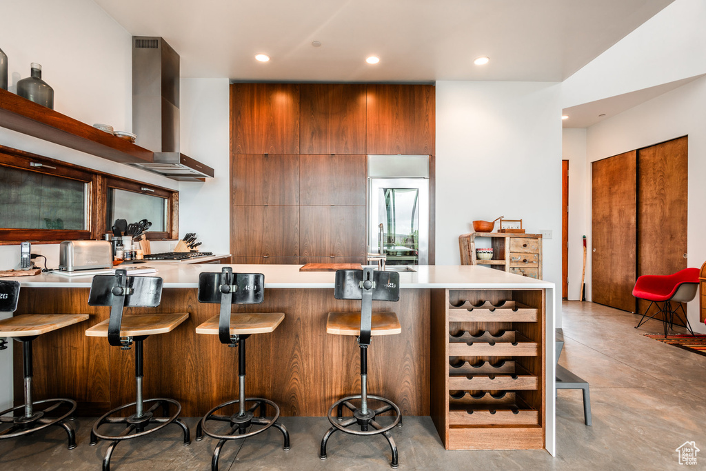 Kitchen featuring wall chimney range hood, stainless steel refrigerator, kitchen peninsula, and a breakfast bar