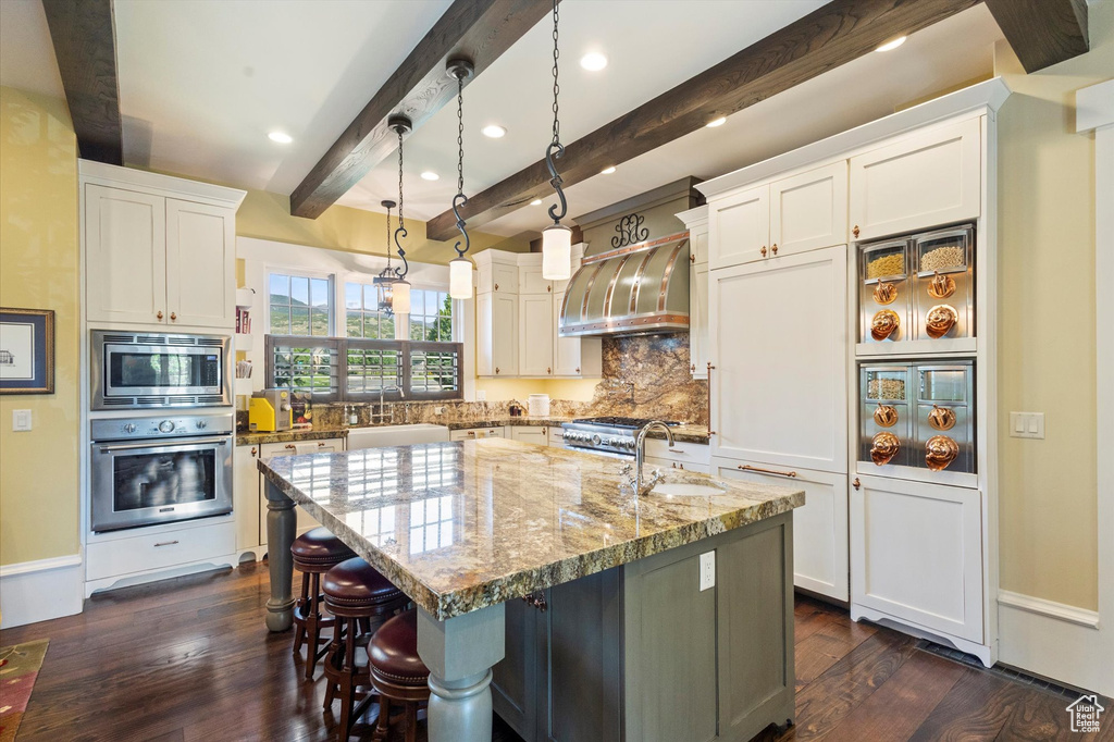 Kitchen featuring dark wood-type flooring, an island with sink, light stone countertops, decorative light fixtures, and stainless steel appliances