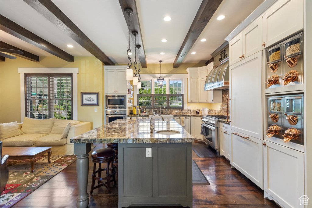 Kitchen featuring decorative light fixtures, appliances with stainless steel finishes, beam ceiling, wall chimney exhaust hood, and light stone countertops