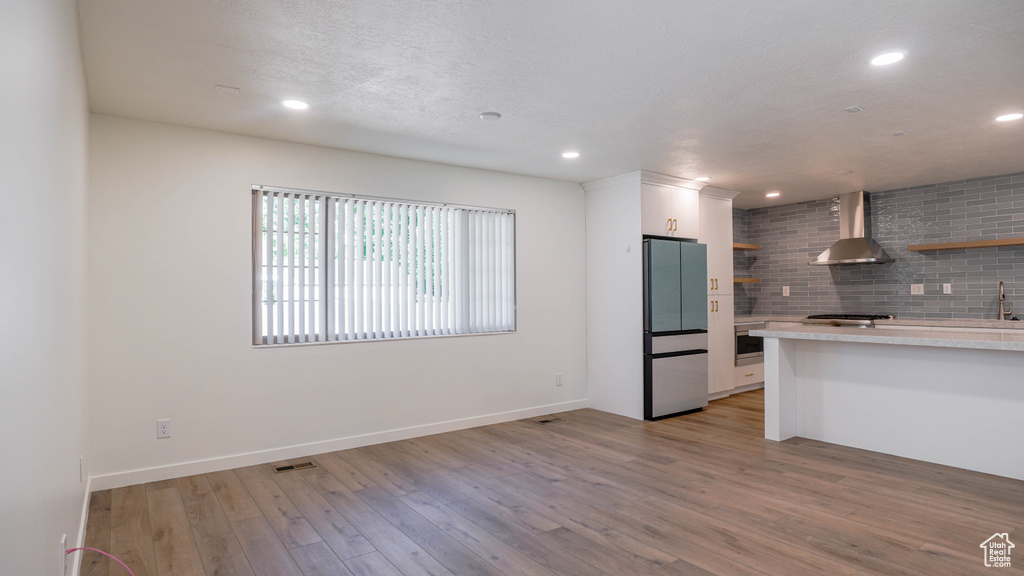 Kitchen with wall chimney range hood, decorative backsplash, stainless steel refrigerator, white cabinetry, and light hardwood / wood-style floors