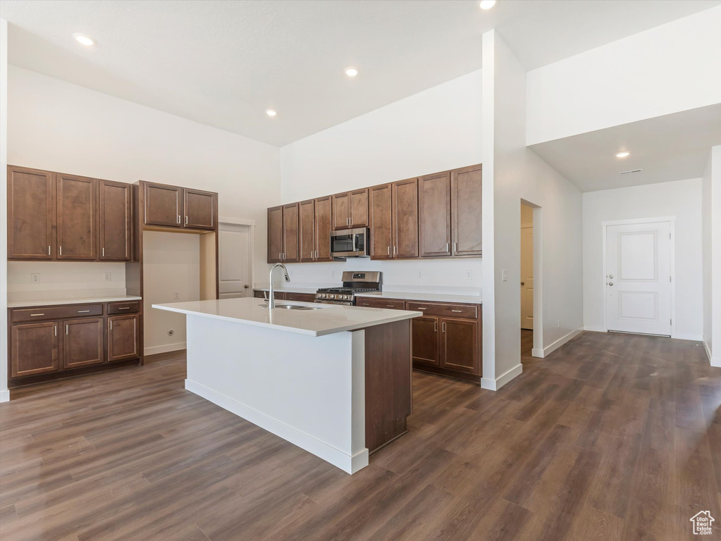 Kitchen with dark hardwood / wood-style floors, a towering ceiling, appliances with stainless steel finishes, sink, and a center island with sink