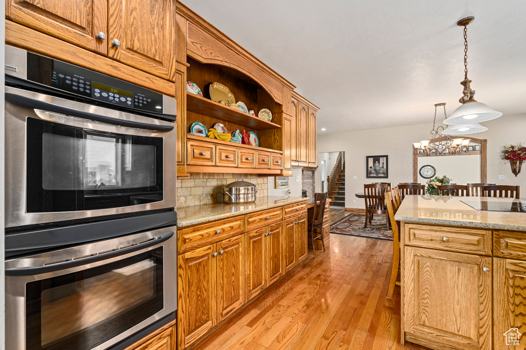 Kitchen with light wood-type flooring, stainless steel double oven, light stone countertops, tasteful backsplash, and hanging light fixtures