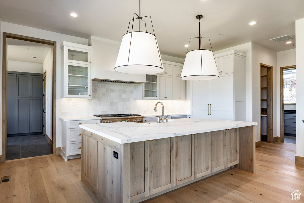 Kitchen with white cabinetry, tasteful backsplash, light stone counters, an island with sink, and light wood-type flooring