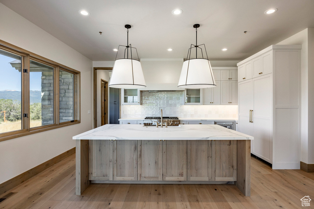 Kitchen with light wood-type flooring, backsplash, a kitchen island with sink, white cabinetry, and light stone countertops