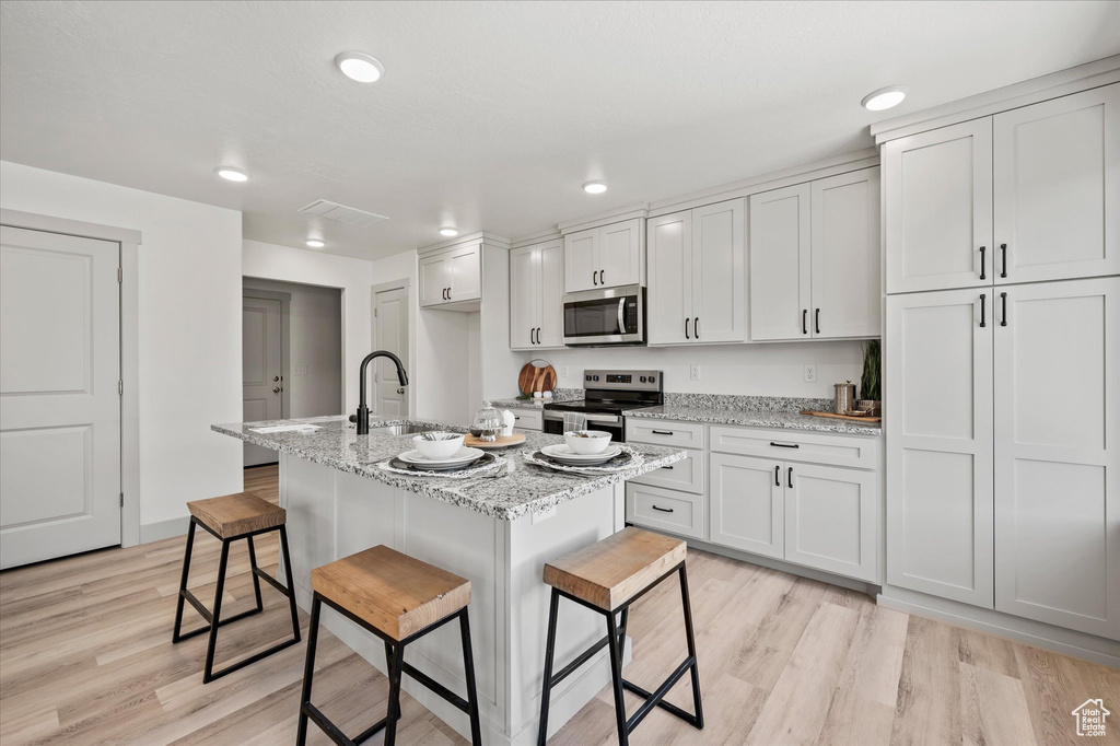 Kitchen featuring stainless steel appliances, light hardwood / wood-style floors, a center island with sink, a breakfast bar, and white cabinets
