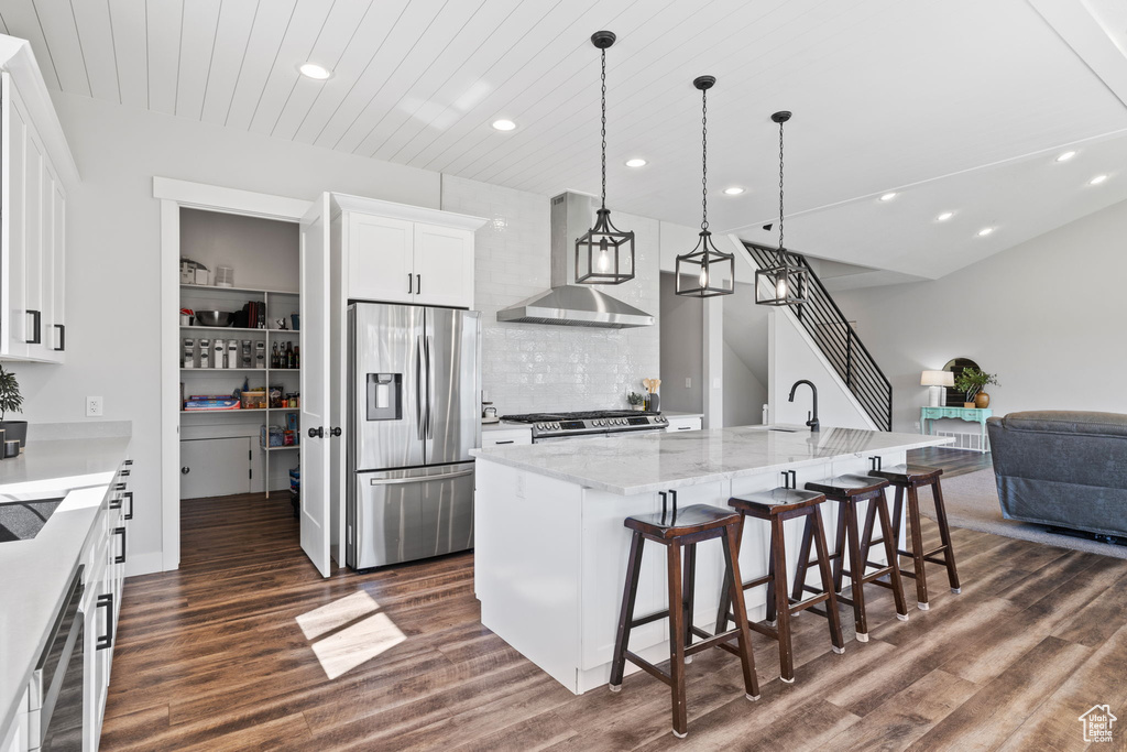 Kitchen featuring stainless steel appliances, a spacious island, wall chimney exhaust hood, dark wood-type flooring, and a kitchen breakfast bar