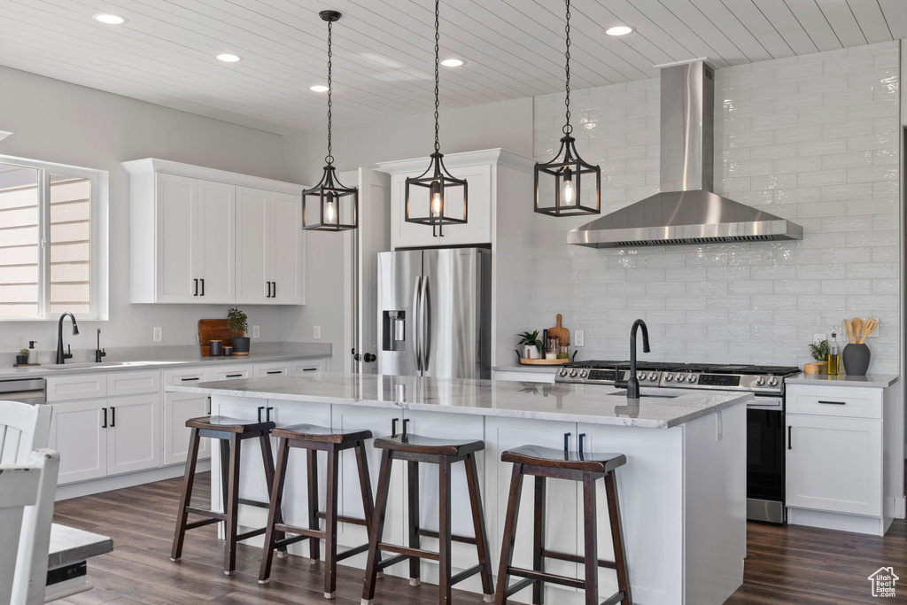 Kitchen with dark hardwood / wood-style floors, light stone countertops, a center island with sink, appliances with stainless steel finishes, and wall chimney range hood