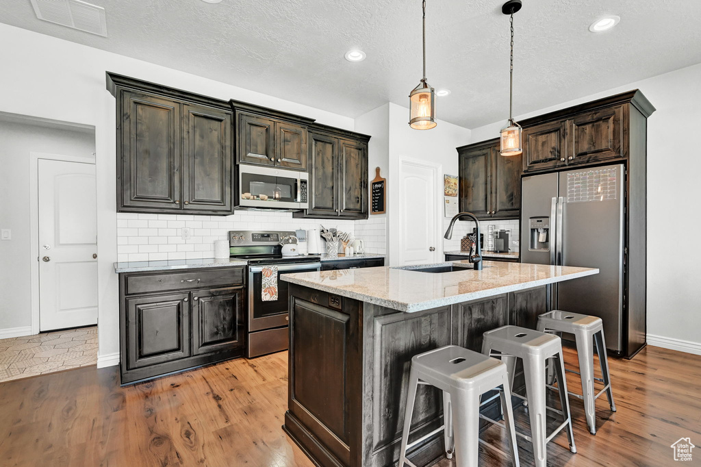 Kitchen featuring backsplash, light wood-type flooring, appliances with stainless steel finishes, light stone countertops, and an island with sink